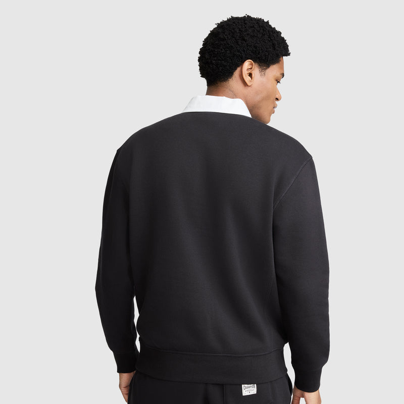 A person with short curly hair is facing away from the camera, wearing black pants and a black Champion Fleece Rugby sweatshirt. The collar of the classic rugby shirt peeks out from under the sweatshirt. The background is plain light gray.