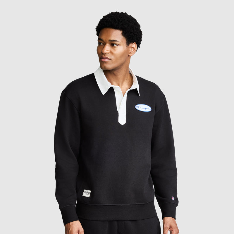 A man wearing the Champion Fleece Rugby shirt in black, featuring a white collar and a small Champion logo on the chest, stands against a plain gray background.