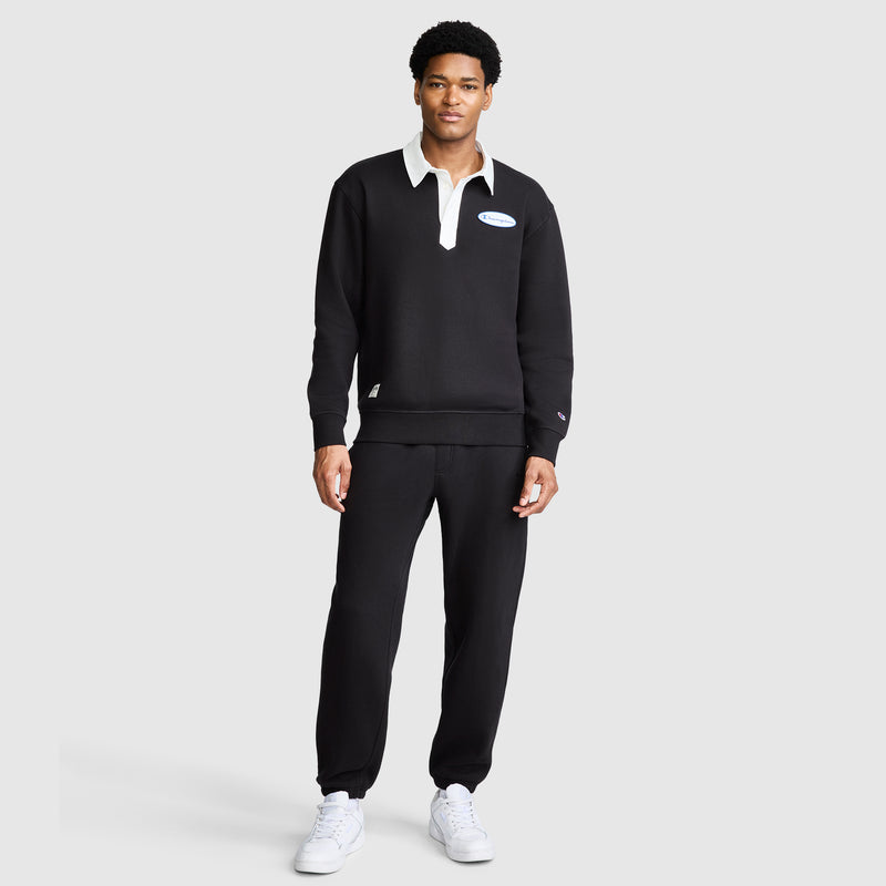 A man stands against a plain background wearing the Champion Fleece Rugby shirt with a white collar, paired with matching black pants and white sneakers. He faces forward with a neutral expression.