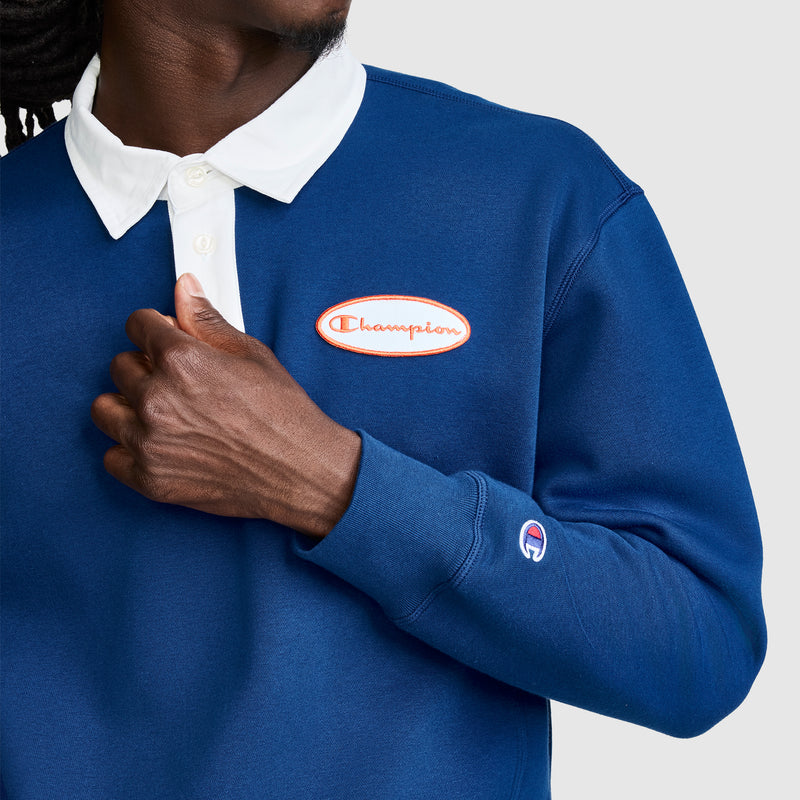 A person wearing the Champion Fleece Rugby shirt in blue with a white collar, holding the collar with one hand. The Champion logo patch is visible on both the chest and sleeve.