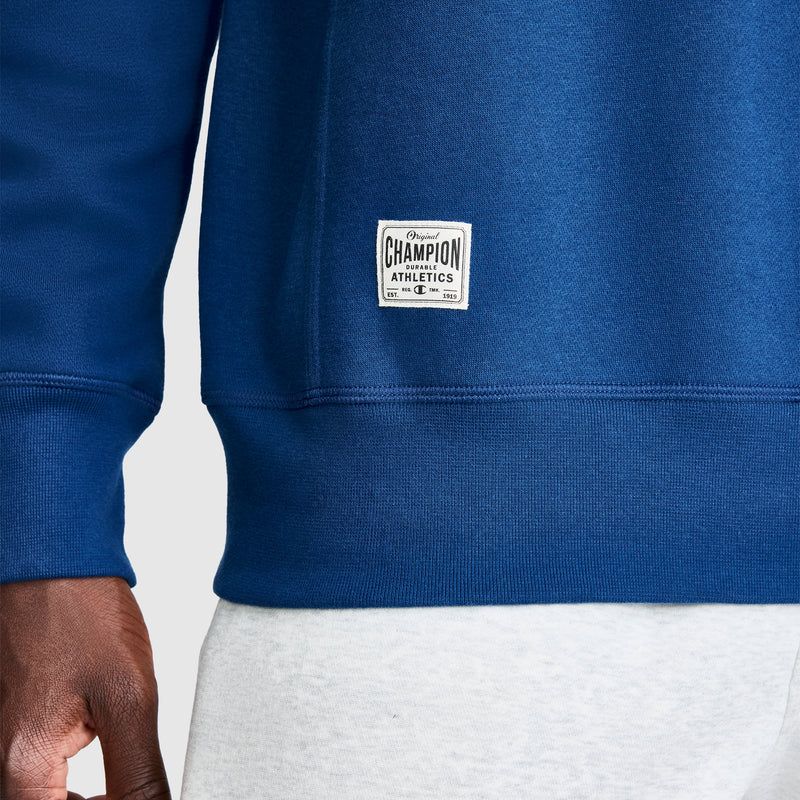 A close-up of a person wearing a blue Champion Fleece Rugby sweatshirt and light gray pants. Only part of the torso and the left hand are visible. The Champion label, sewn near the hem, highlights this classic Fleece Rugby style.