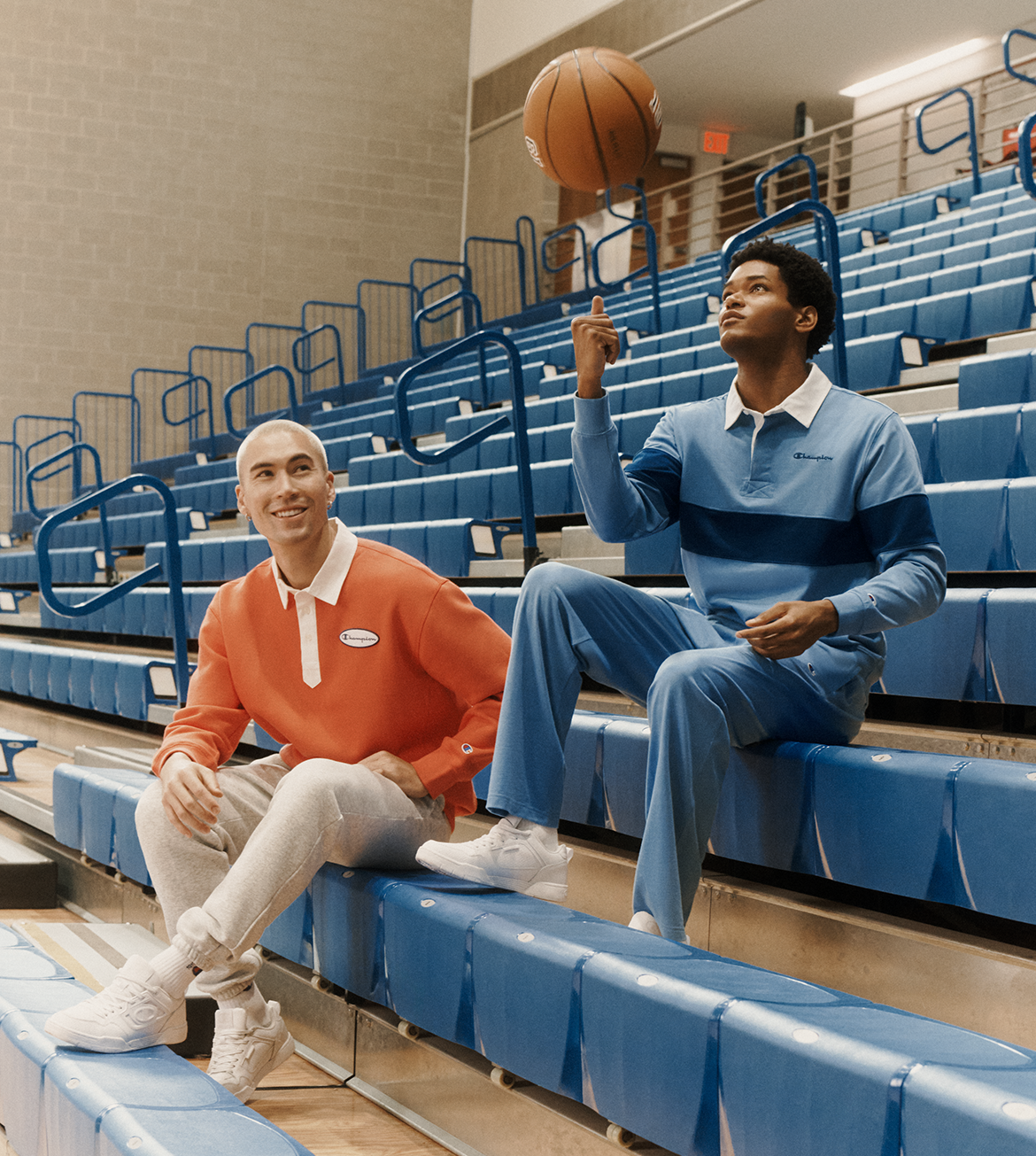 Two men sitting on blue gym bleachers in an indoor basketball court. One man wears an orange long-sleeve polo with white pants and sneakers, while the other wears a blue and navy striped long-sleeve polo with matching blue pants and white sneakers.
