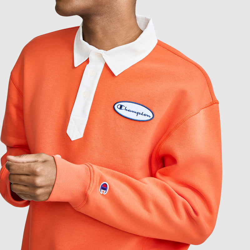 A person wearing a Champion Fleece Rugby shirt in orange, featuring a white collar and embroidered Champion logos on the chest and sleeve. Their face is partially out of frame.