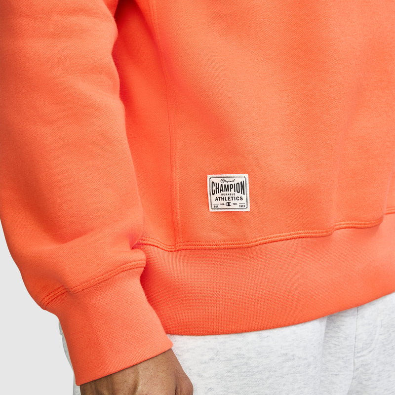 A person wears a bright orange Champion Fleece Rugby shirt and light gray pants. Only the torso and arm are visible, with a Champion logo label sewn near the hem of the shirt.