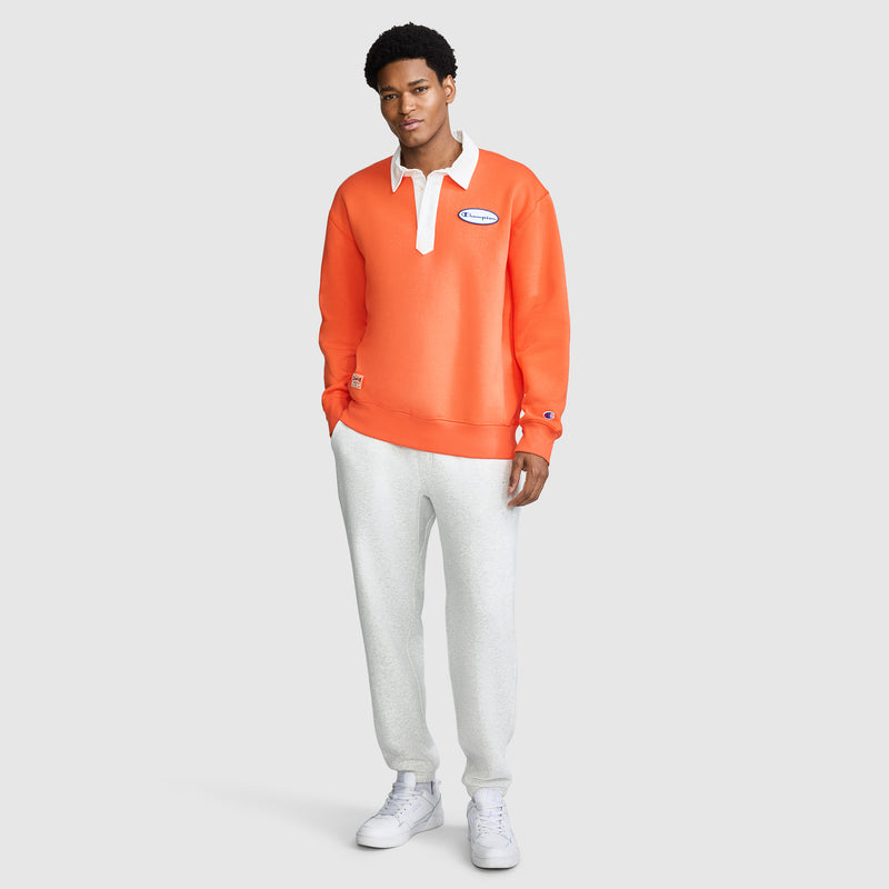A man stands against a light background, wearing the Champion Fleece Rugby—a men's orange pullover with a white collar and patch—paired with light gray sweatpants and white sneakers. He faces forward with a relaxed expression.