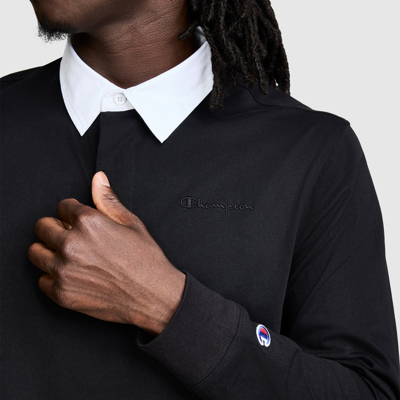 A person in a black Champion Long Sleeve Rugby Shirt with Twill Collar, featuring the embroidered C logo on the chest and sleeve, gives a thumbs up. The shirt’s white collar stands out, and the person’s face is partially out of frame.