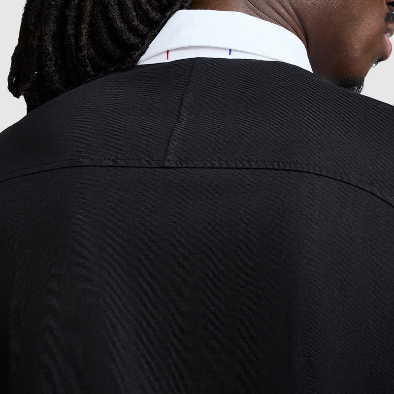 Close-up of the back of a person wearing the Champion Long Sleeve Rugby Shirt with Twill Collar and an embroidered C logo. Only part of their head, neck, and shoulders is visible. The shirt features a black body, white twill collar, and relaxed fit—and the person has dark skin and short dreadlocks.