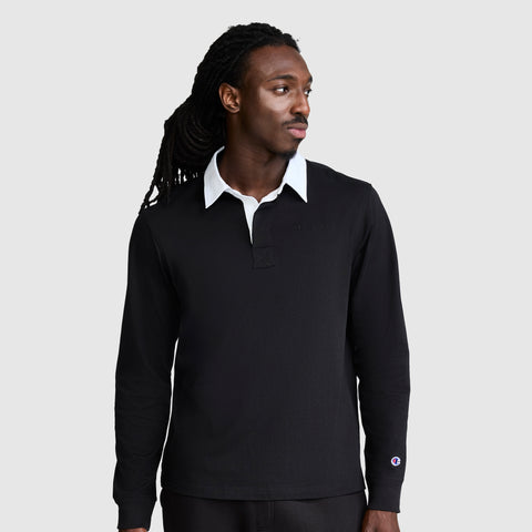 A man with long, dark hair wears the Champion Long Sleeve Rugby Shirt with Twill Collar and Embroidered C Logo, featuring a black body and white collar. He looks slightly to his left against a plain, light gray background.