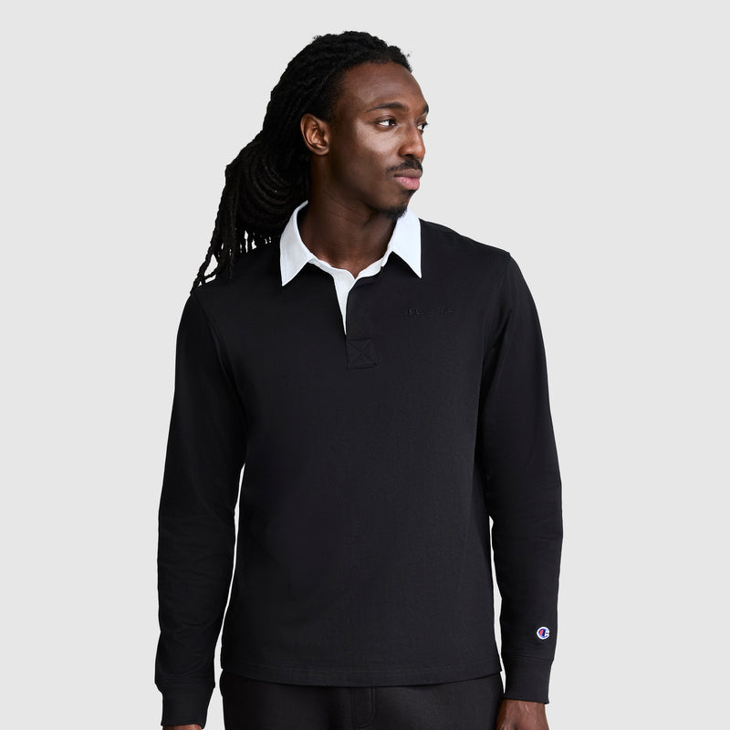 A man with long, dark hair wears the Champion Long Sleeve Rugby Shirt with Twill Collar and Embroidered C Logo, featuring a black body and white collar. He looks slightly to his left against a plain, light gray background.