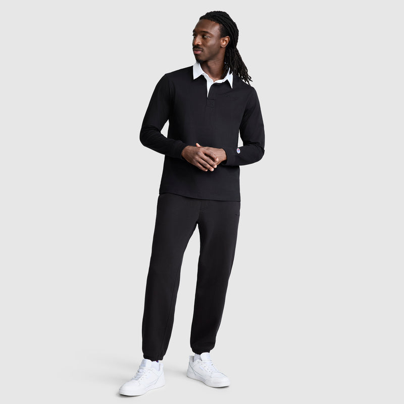 A man with long dreadlocks stands against a light gray background, wearing a black Champion Long Sleeve Rugby Shirt with a twill collar and embroidered C logo over a white undershirt, paired with black jogger pants and white sneakers. He looks to his left in a relaxed pose.