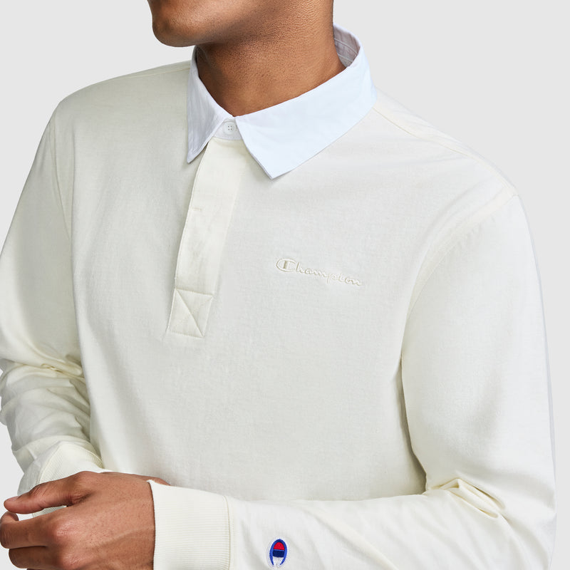A person wearing the Champion Long Sleeve Rugby Shirt with Twill Collar and embroidered C logo, featuring cream-colored fabric and a classic white collar. The logos are visible on the chest and sleeve, while the person’s face is partially out of frame.