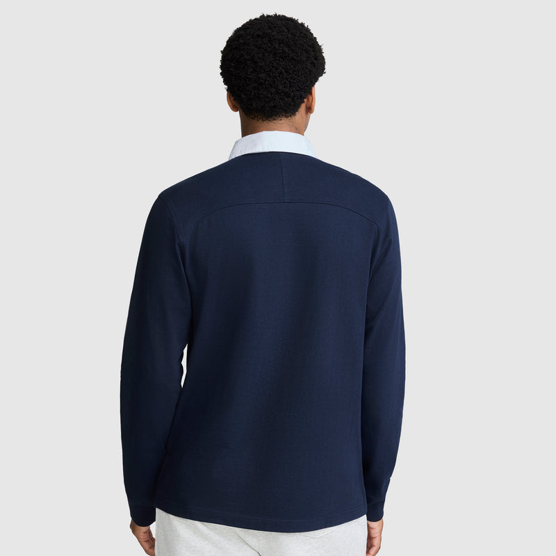 A person with short curly hair is shown from the back, wearing a Champion Long Sleeve Rugby Shirt with Twill Collar and Embroidered C Logo, layered over a white collared shirt and paired with light-colored pants, standing against a plain light gray background.