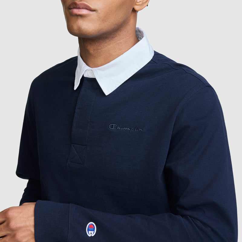 A person wearing a navy blue Champion Long Sleeve Rugby Shirt with Twill Collar, featuring the embroidered C logo on the chest and sleeve. The background is plain light gray.