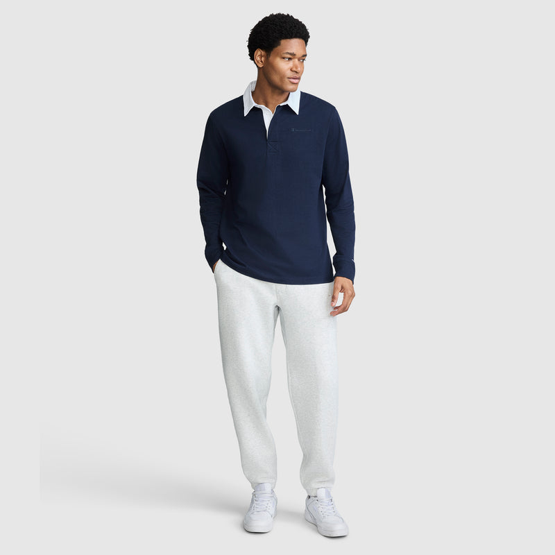 A man is wearing a navy Champion Long Sleeve Rugby Shirt with Twill Collar and Embroidered C Logo, paired with light gray sweatpants and white sneakers, standing and looking to the side against a plain light gray background.
