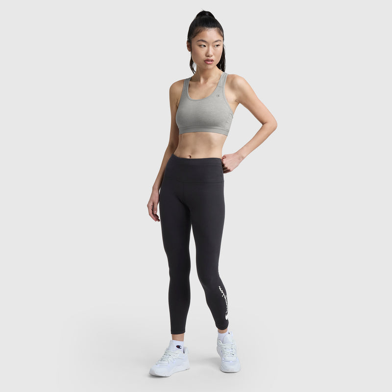 A woman stands against a light gray background, wearing the Champion Compression Sports Bra with C Logo, high-waisted black leggings, and white athletic shoes. She poses with one hand on her hip, gazing slightly to the side.