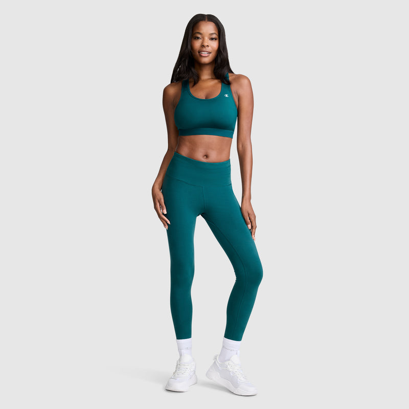 A woman wearing a moisture-wicking teal Compression Sports Bra, C Logo by Champion with matching leggings, white socks, and sneakers stands smiling against a plain light gray background.