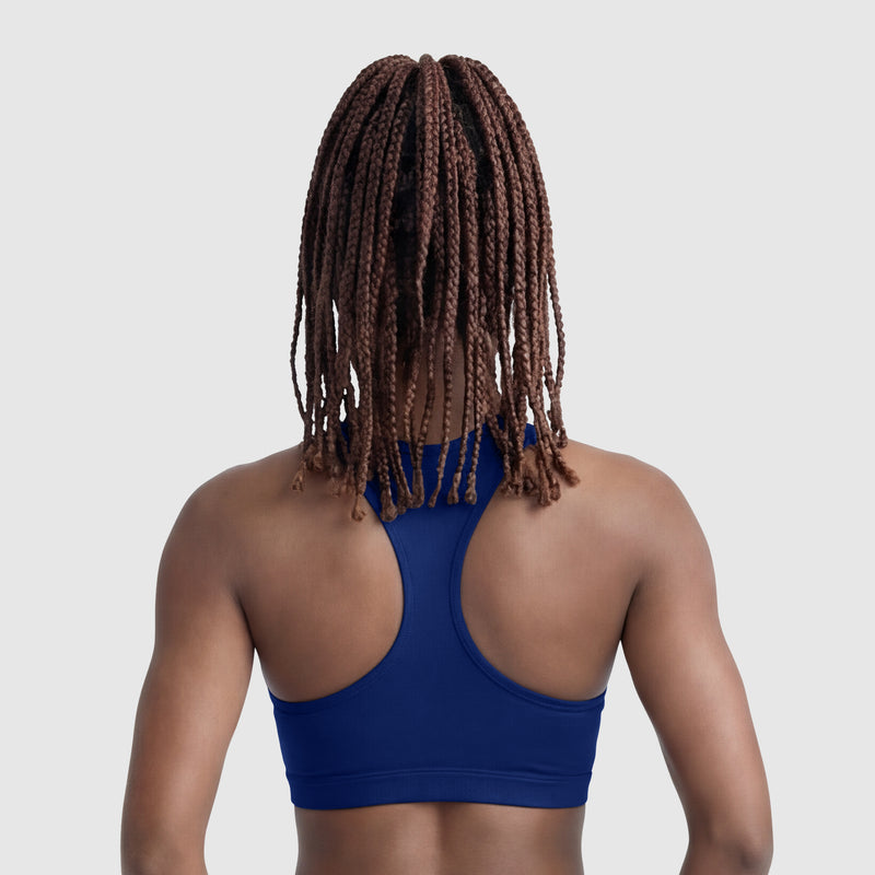A person with long, brown box braids is seen from behind, wearing a blue Champion Compression Sports Bra with C Logo, offering moderate support against a plain light gray background.