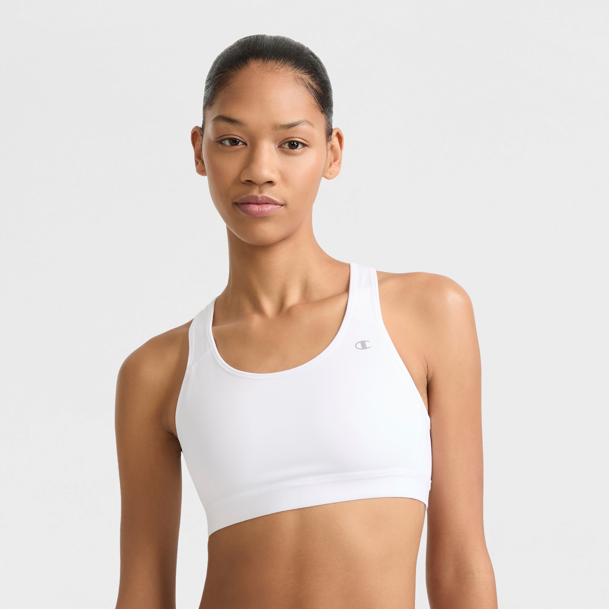 A woman with her hair tied back wears a Champion Compression Sports Bra featuring moderate support and a small C Logo on the left, standing against a plain light gray background.