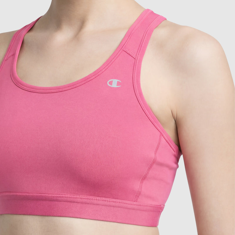 A woman wearing the Champion Compression Sports Bra in pink with a racerback and small white C logo, offering moderate support and moisture-wicking, poses against a plain light gray background.