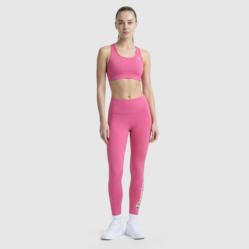 A woman faces forward against a light gray background, wearing Champion's Compression Sports Bra, C Logo, and matching pink leggings with white sneakers.