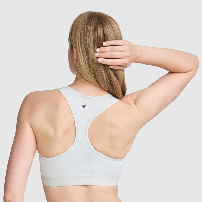 A person with long blonde hair faces away, wearing the Champion Compression Sports Bra featuring a racerback design and the iconic C logo. One arm is raised with their hand touching the back of their head. The moisture-wicking fabric offers moderate support, while the plain gray background keeps attention on the sleek activewear.