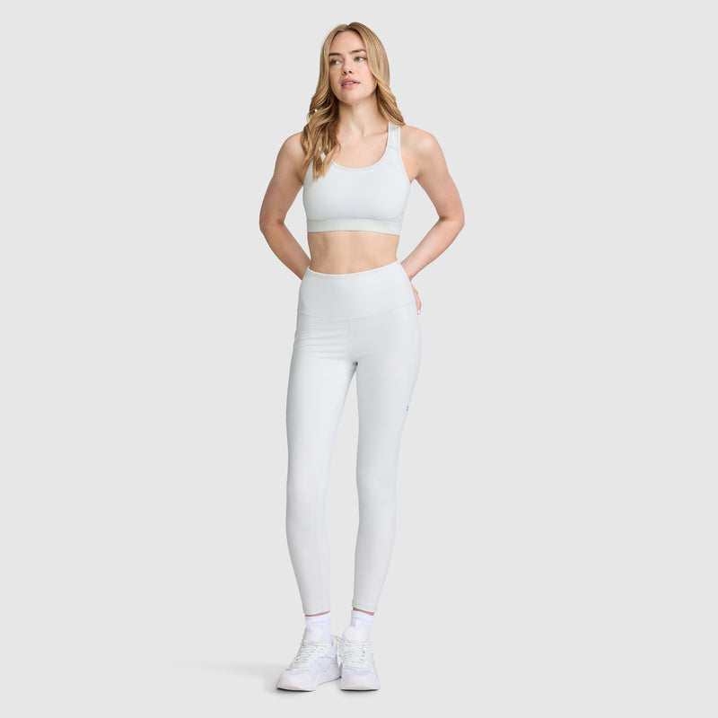 A woman wearing a white Champion Compression Sports Bra with the C Logo, paired with white high-waisted leggings and white sneakers, stands against a plain light gray background, facing forward with her hands resting on her hips.