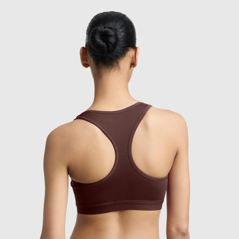 A woman with dark hair in a bun is seen from the back, wearing the Champion Compression Sports Bra, C Logo in maroon with moderate support against a plain light gray background.