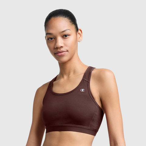 A woman with pulled-back hair wearing a brown Champion Compression Sports Bra, C Logo, stands against a plain light gray background, smiling softly at the camera. This bra features moisture-wicking fabric and provides moderate support for everyday comfort.