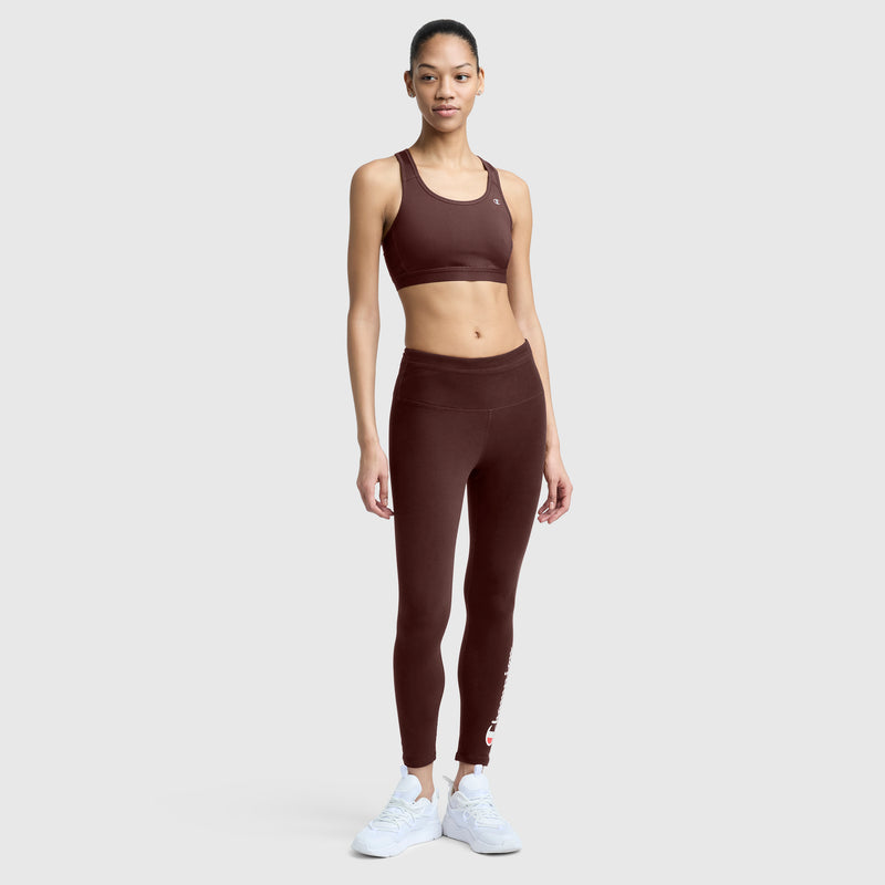 A woman stands facing forward, wearing a matching maroon Champion Compression Sports Bra with C Logo and leggings set, paired with white sneakers. She is on a plain light gray background.