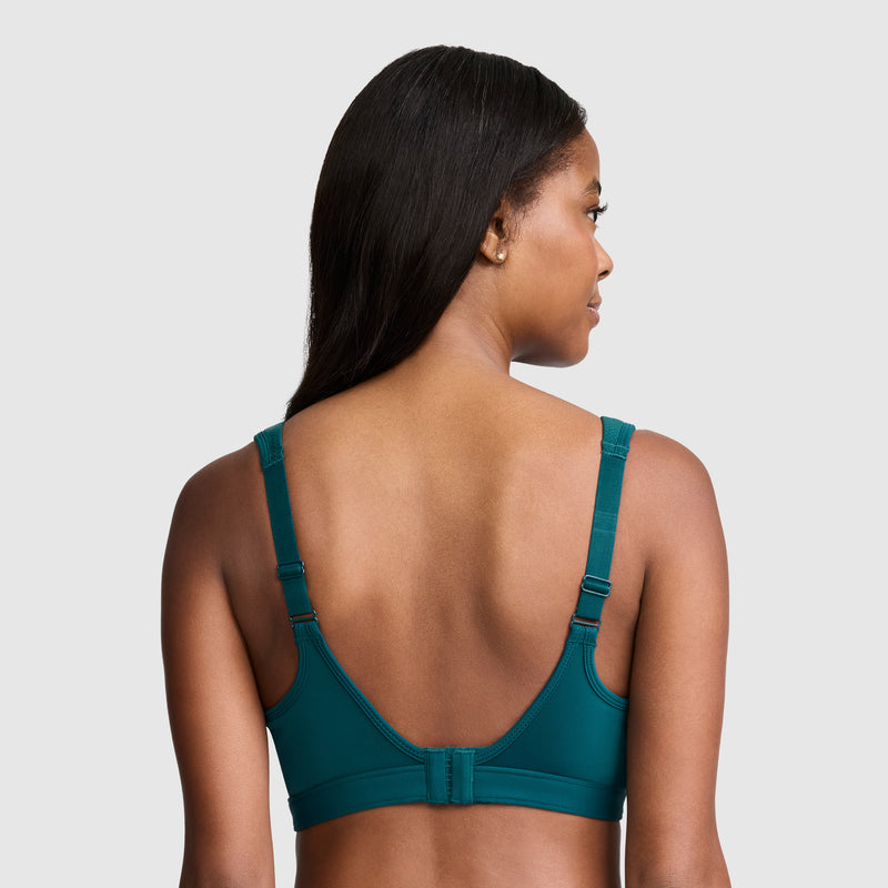 A woman with long dark hair, shown from the back against a plain light gray background, models the Champion Spot Comfort High-Impact Sports Bra, C Logo in teal, featuring adjustable straps and a hook-and-eye closure.