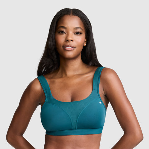 A woman with long dark hair wears the Champion Spot Comfort High-Impact Sports Bra, featuring the C logo in teal for maximum support, and smiles slightly while standing against a plain, light gray background.