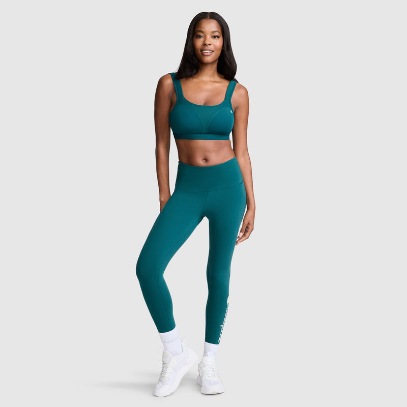 A woman stands and smiles, wearing the Champion Spot Comfort High-Impact Sports Bra, C Logo in teal for maximum support, paired with matching leggings, white socks, and white athletic shoes against a plain light gray background. The moisture-wicking fabric of the sports bra keeps her cool and comfortable during any activity.