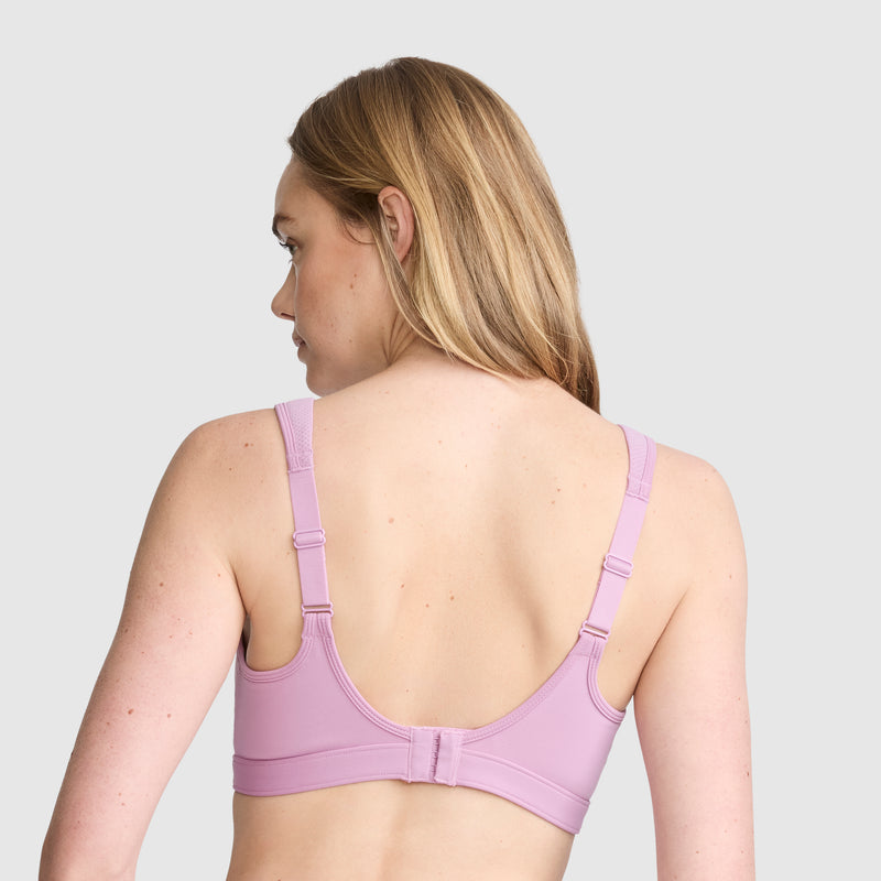 A woman with long blonde hair is shown from the back, wearing the Champion Spot Comfort High-Impact Sports Bra, C Logo in light purple with adjustable straps designed for maximum support against a plain light gray background.