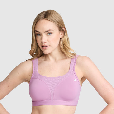 A woman with light skin and blonde hair models the Champion Spot Comfort High-Impact Sports Bra, C Logo, in lavender. Featuring wide straps for maximum support and moisture-wicking comfort, she stands against a plain light gray background, gazing slightly to the side with a neutral expression.