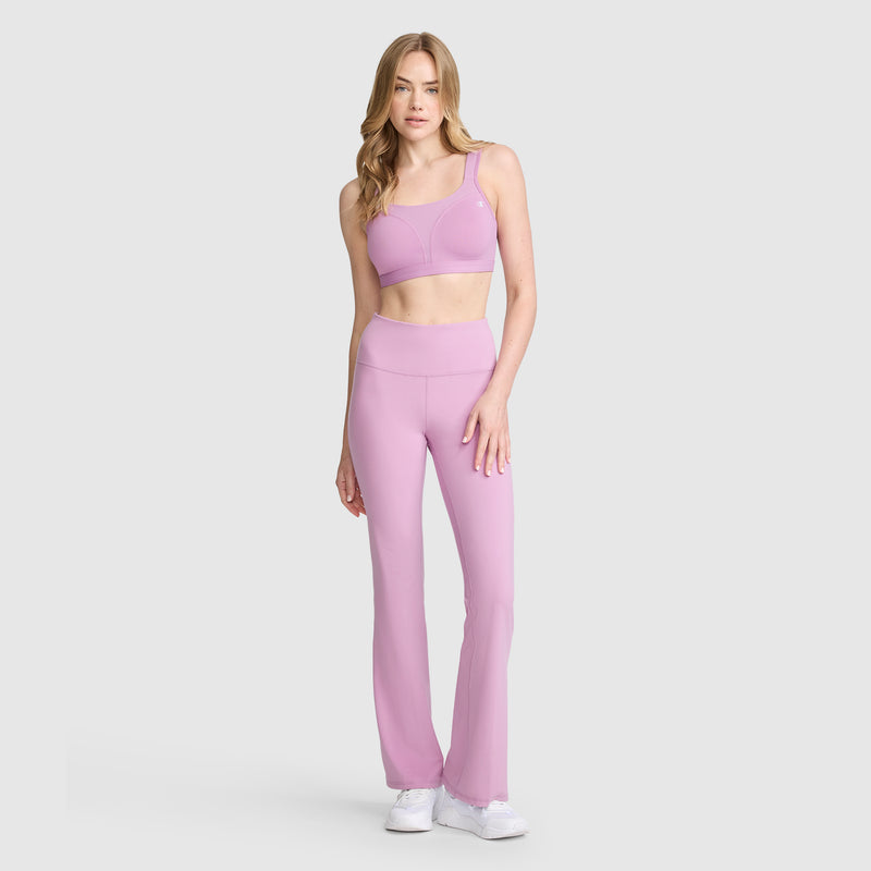 A woman is wearing the Champion Spot Comfort High-Impact Sports Bra, C Logo in light purple, paired with matching high-waisted flare leggings. She stands with one hand by her side and the other resting on her thigh, showcasing a look designed for maximum support. The set is completed with white sneakers against a plain light background.
