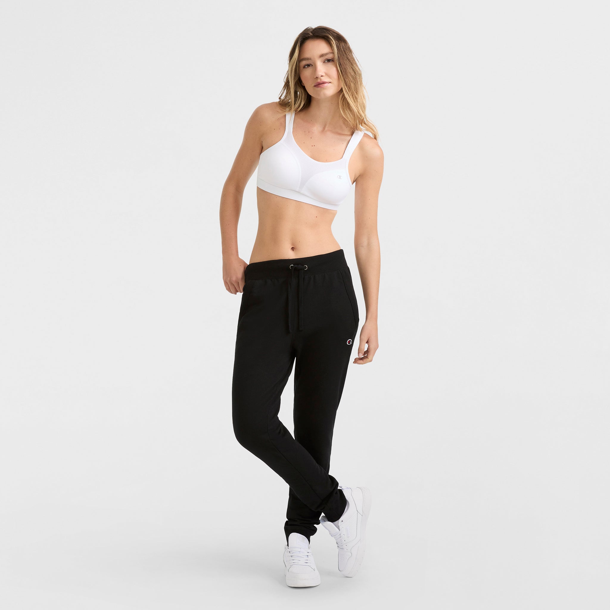 A woman poses against a plain background wearing the Champion Spot Comfort High-Impact Sports Bra, black drawstring jogger sweatpants, and white sneakers. She stands with one leg crossed over the other, left hand on her hip.