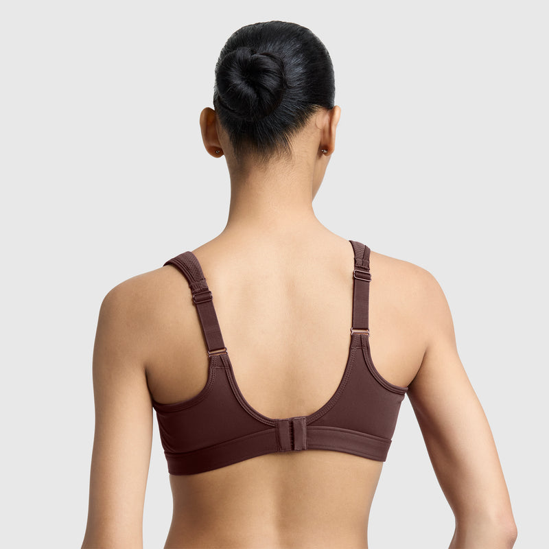 A person with dark hair in a bun is shown from the back, wearing the Champion Spot Comfort High-Impact Sports Bra, C Logo—a brown high-impact sports bra featuring wide straps and an adjustable hook closure—set against a plain light gray background.