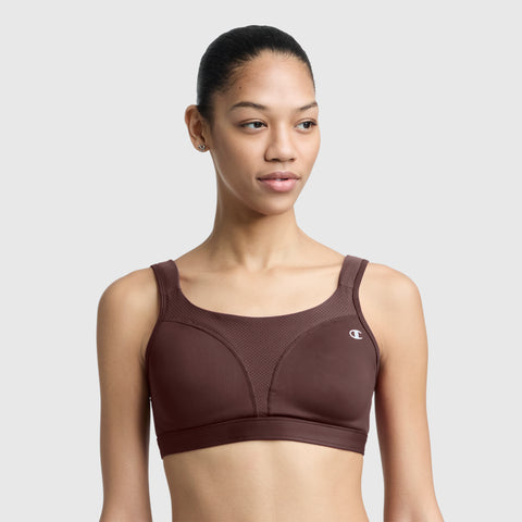 A woman with tied-back hair wears the Champion Spot Comfort High-Impact Sports Bra featuring the C Logo in brown, standing against a plain light gray background and looking slightly to the side.