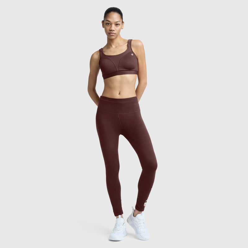 A woman stands facing forward against a plain light gray background, wearing the Champion Spot Comfort High-Impact Sports Bra with the C Logo in maroon, paired with matching leggings and white sneakers.