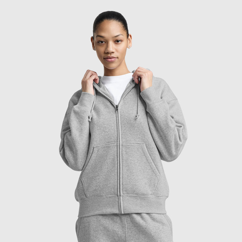 A woman faces forward against a plain light background, wearing the Champion Powerblend Fleece Zip-Up Hoodie, C Logo in light gray over a white shirt and matching sweatpants, holding the hoodie collar with both hands.