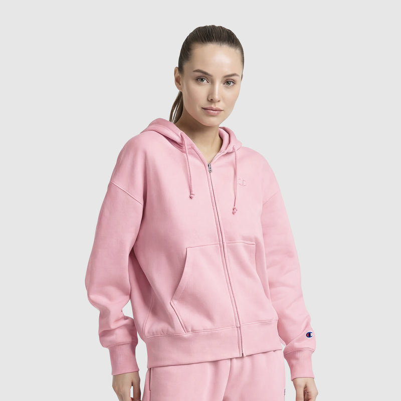 A woman with light skin and brown hair pulled back stands against a plain light background, wearing a light pink Champion Powerblend Fleece Zip-Up Hoodie, C Logo, with front pockets and hood, paired with matching pink pants.