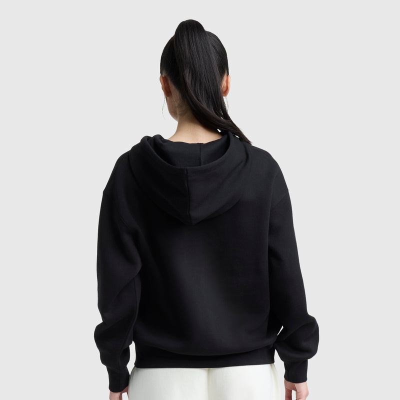 A person with long dark hair in a ponytail wears the Champion Powerblend Fleece Zip-Up Hoodie, C Logo in black with white pants, standing with their back to the camera against a plain light gray background.