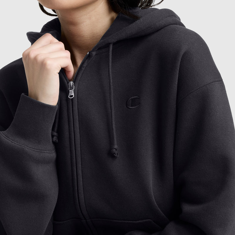 A person wears a dark gray Champion Powerblend Fleece Zip-Up Hoodie with a hood, drawstrings, and the C logo stitched on the chest, holding the zipper near their chin. The background is plain and light-colored.
