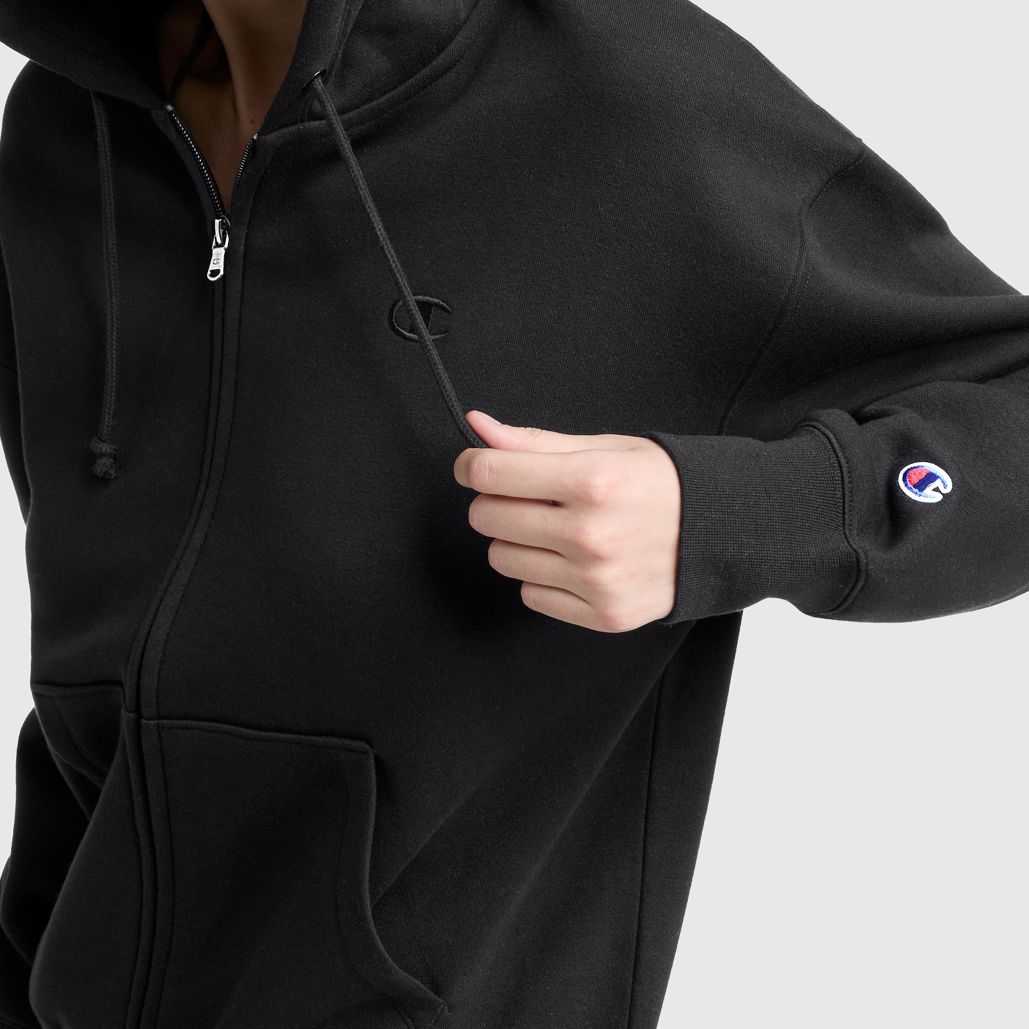 A person wears a black Champion Powerblend Fleece Zip-Up Hoodie, C Logo, pulling the drawstring with one hand. Champion branding is visible on the chest and sleeve against a plain gray background.