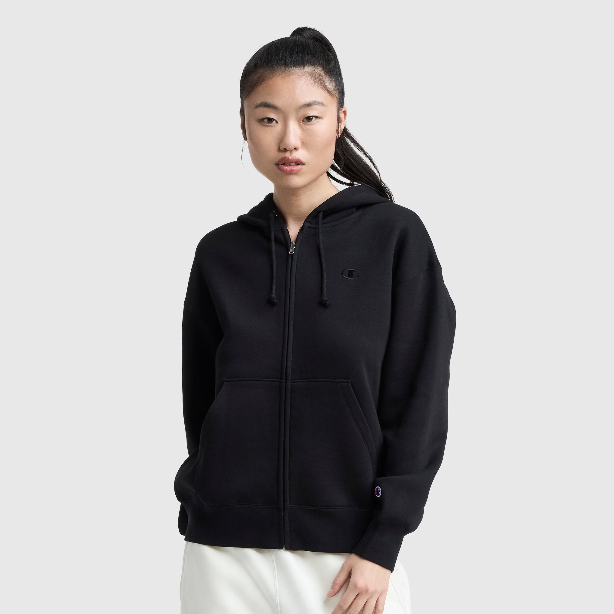 A young woman with long black hair in a ponytail wears the Champion Powerblend Fleece Zip-Up Hoodie, C Logo, with front pockets and white pants, standing against a plain light gray background.