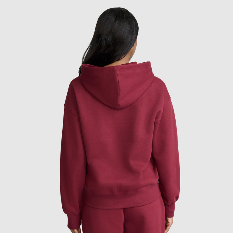 A person with long dark hair, seen from behind, wears a matching maroon Champion Powerblend Fleece Zip-Up Hoodie with the C Logo and sweatpants set. The hood is down, and the background is plain light gray.