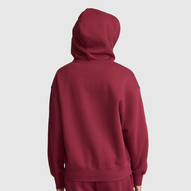 A person facing away from the camera, wearing a maroon Champion Powerblend Fleece Zip-Up Hoodie, C Logo, with the hood up and matching maroon pants, against a plain light gray background.