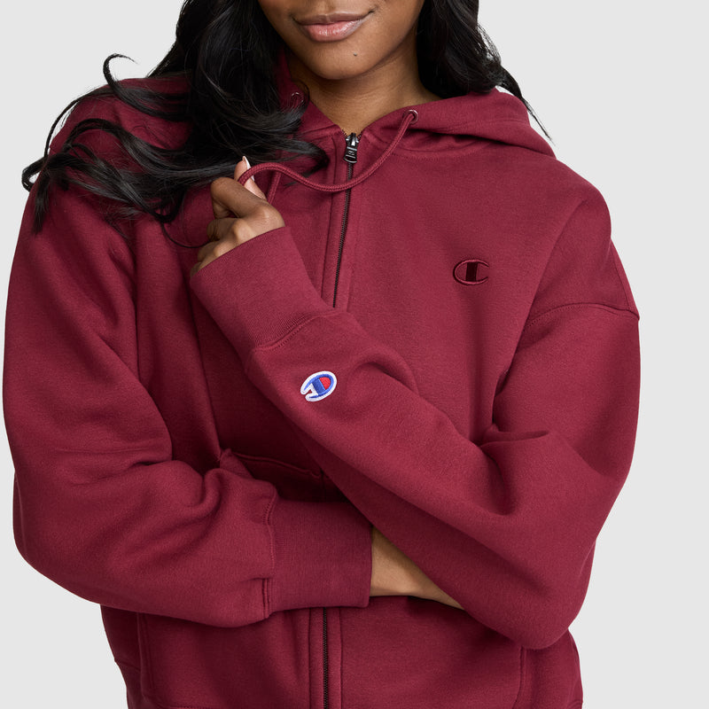 A woman wearing a maroon Champion Powerblend Fleece Zip-Up Hoodie, C Logo, holds the collar with one hand. The Champion logo is visible on both the chest and the sleeve. She stands against a plain, light-colored background.