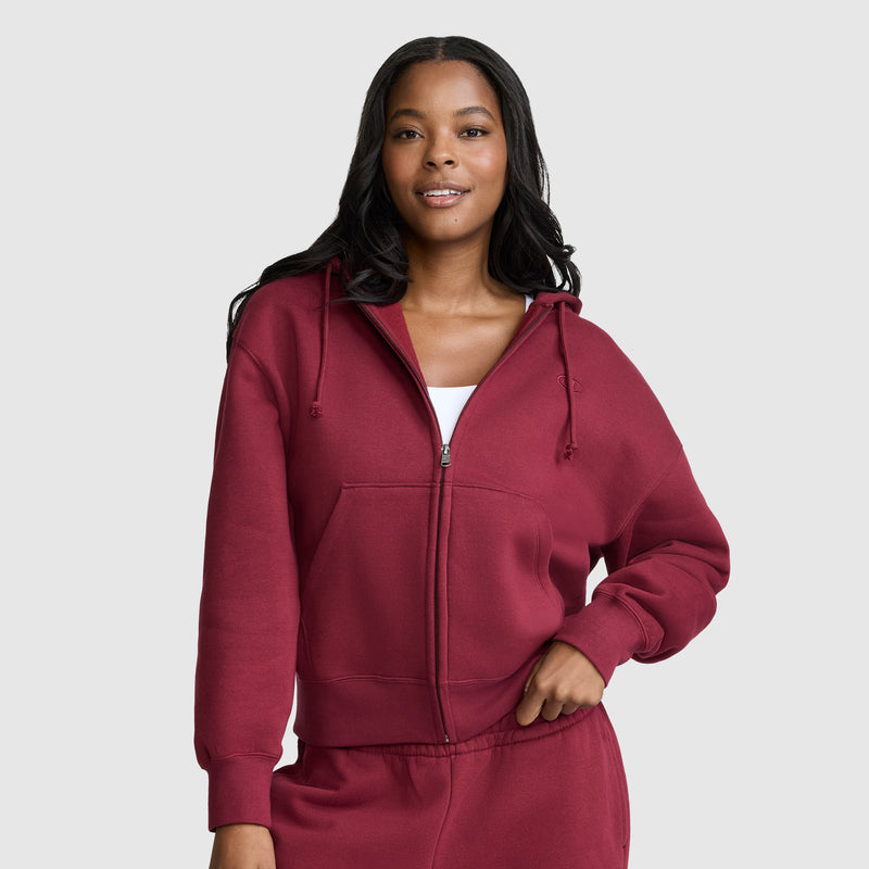 A woman wearing a maroon Champion Powerblend Fleece Zip-Up Hoodie, C Logo, and matching sweatpants poses confidently with one hand on her hip against a plain light gray background.