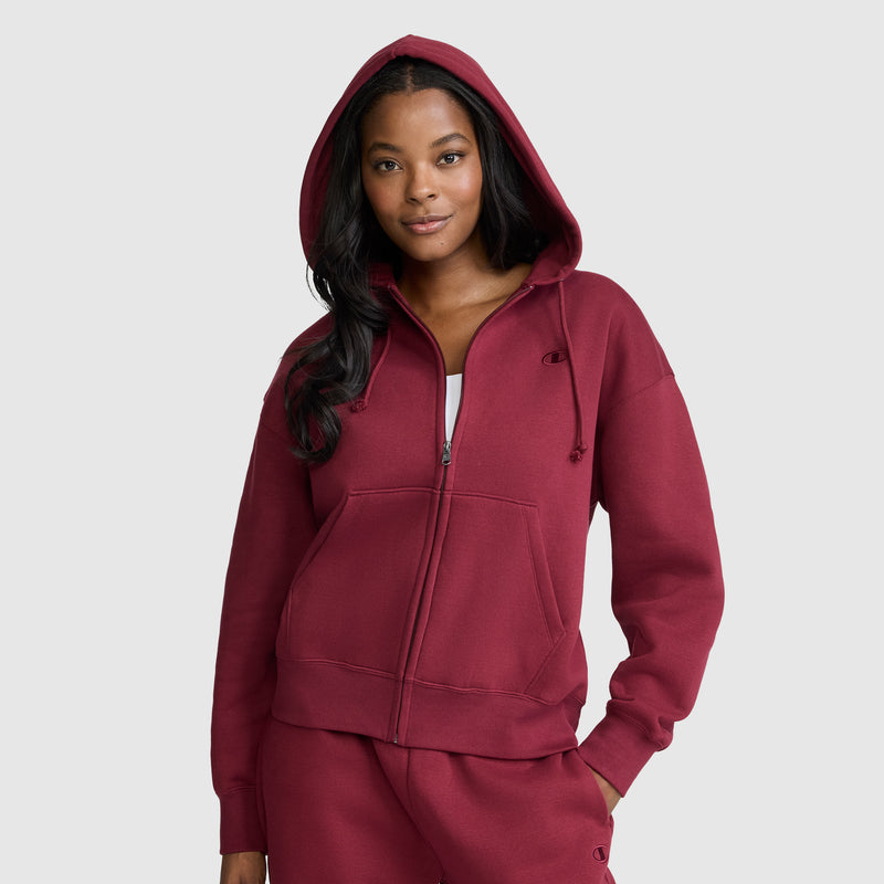 A woman wearing a maroon Champion Powerblend Fleece Zip-Up Hoodie, C Logo, with the hood up, stands with one hand in her pocket and a slight smile against a plain light gray background. This Champion hoodie blends comfort and style for a relaxed, athletic look.